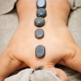 From above of topless female client with hot stones on back lying on bed during massage session in spa salon